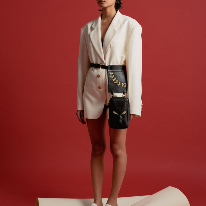 Woman in white blazer dress with black leather belt bag and white slip-on shoes against red backdrop