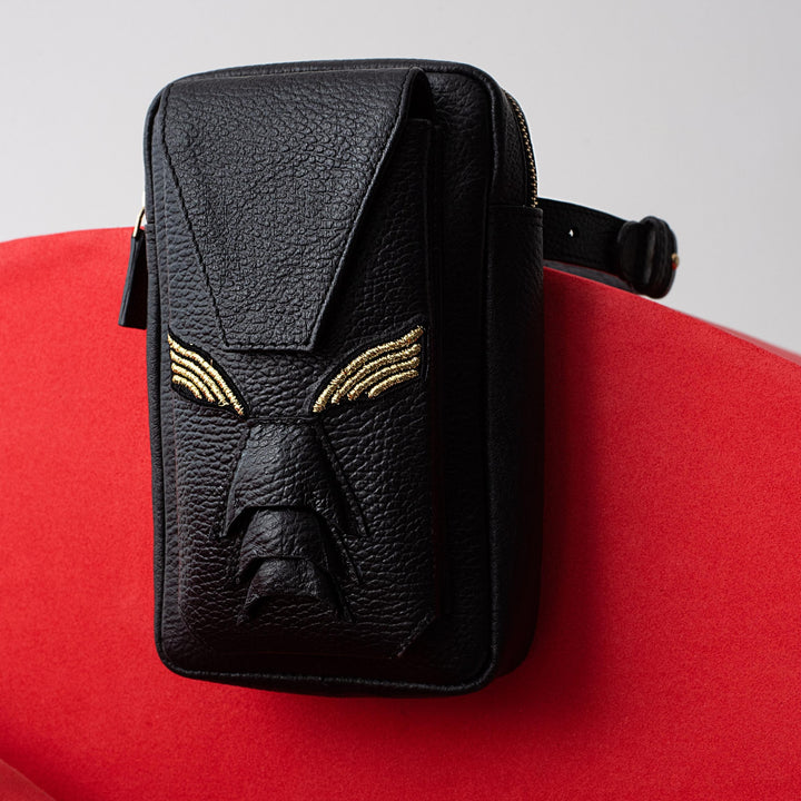 Black textured leather belt bag with gold embroidered detail resembling a face on red background