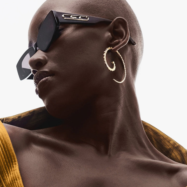 Close-up of stylish bald woman wearing black sunglasses, gold spiral hoop earring, and mustard yellow top