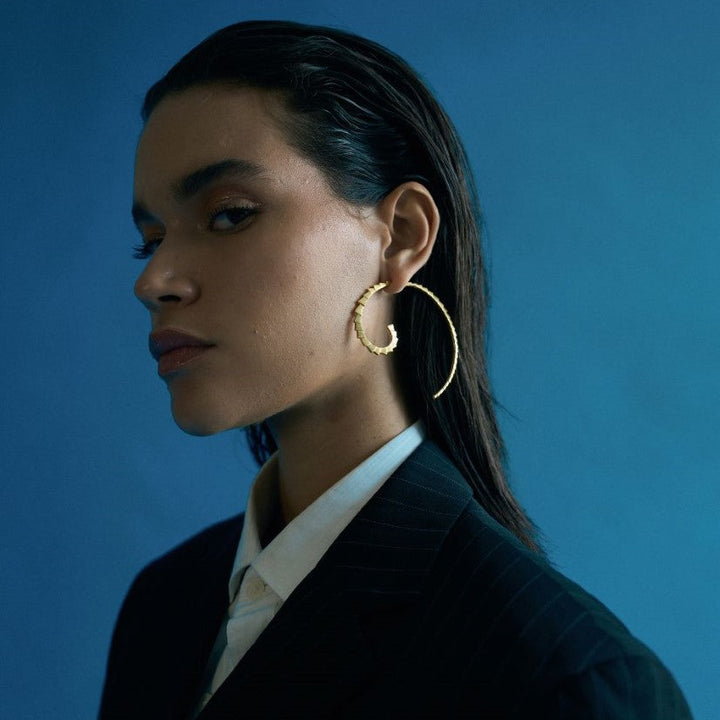 Profile of a woman wearing large gold spiral hoop earrings against a plain blue background