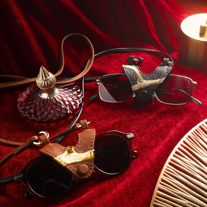 Two pairs of glasses with leather holders on textured red velvet, next to a vintage glass perfume bottle and a brass lamp