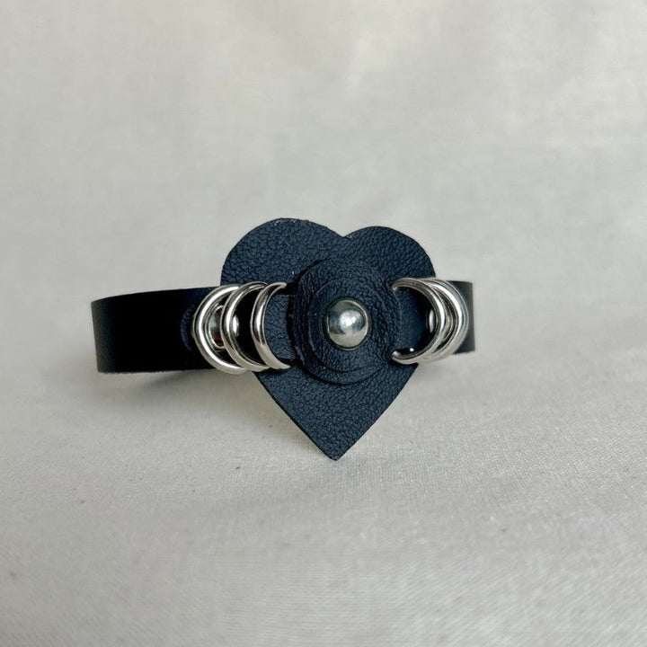 Black leather cuff bracelet with heart-shaped centerpiece and silver ring studs on neutral background
