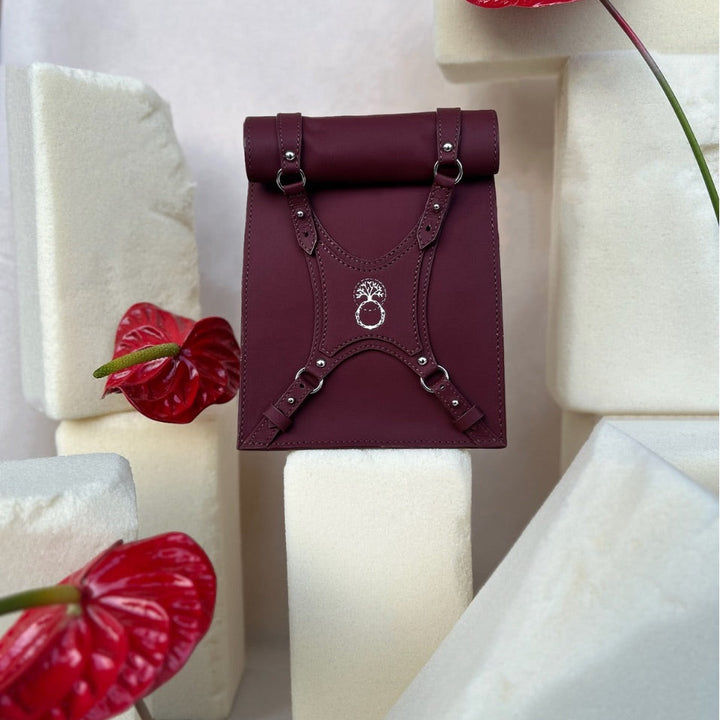 Maroon leather roll-top backpack with silver buckles, displayed on white foam blocks with red anthurium flowers