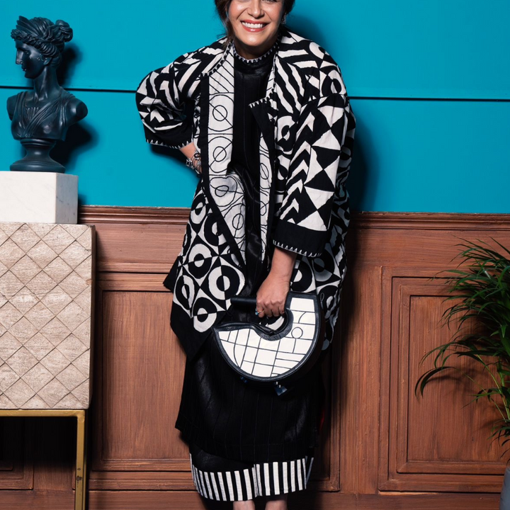 Smiling woman in black and white geometric print outfit holding matching handbag, standing by blue wall