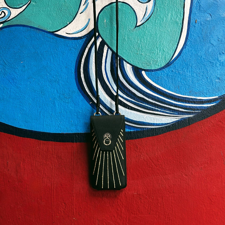 Black leather phone sling bag with gold embroidery hanging against colorful blue, green, red wall mural