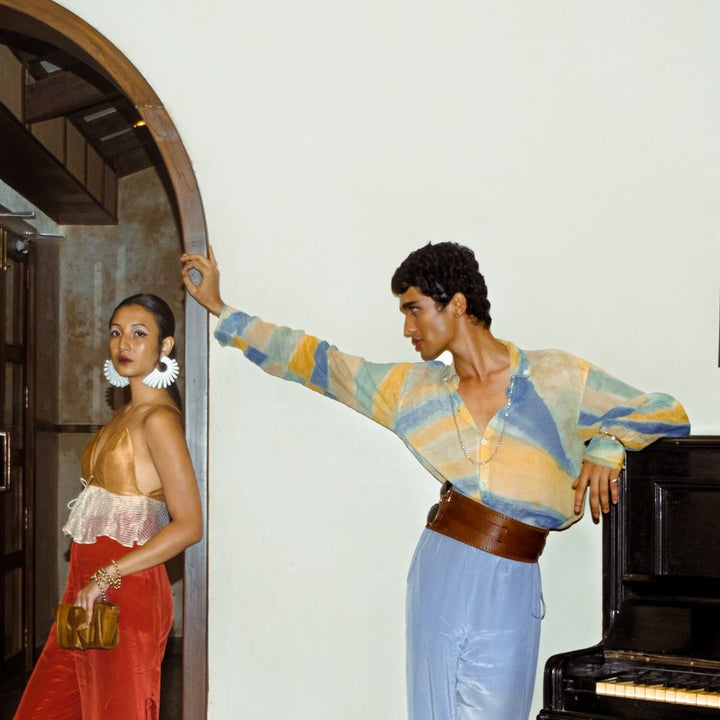 Fashionable man in pastel tie-dye shirt and wide tan leather waist belt posing by piano, woman in orange pants and gold top nearby