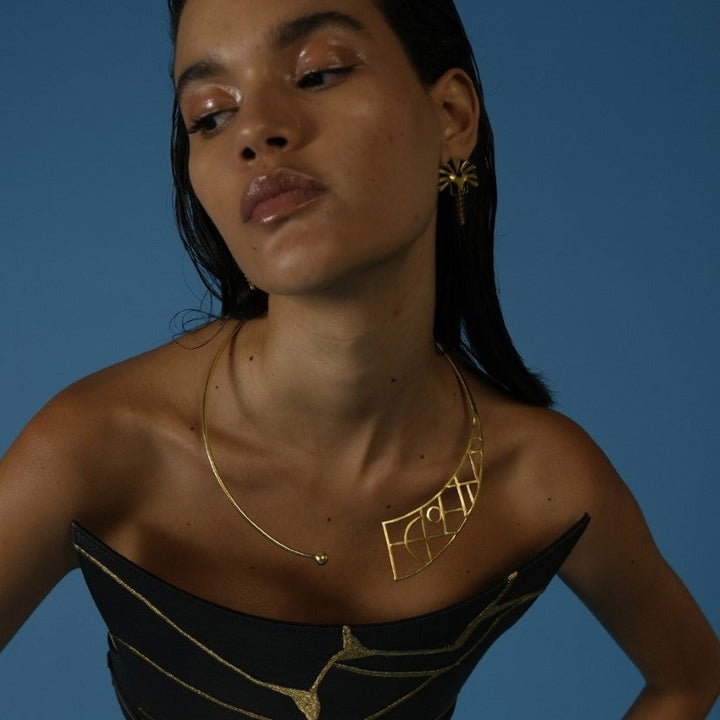 Woman wearing a sculptural metal wire choker with geometric design and matching gold earrings against blue background