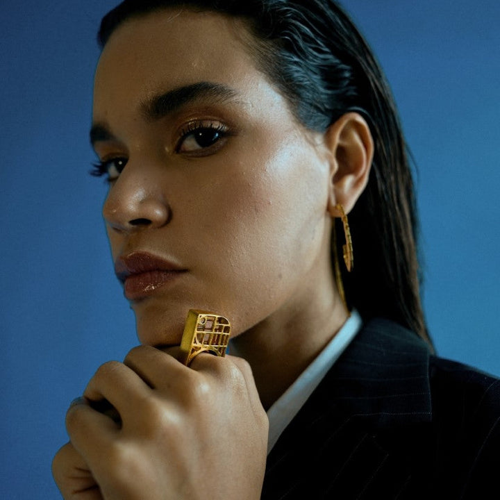 Close-up of woman wearing geometric gold statement ring and matching earrings against blue background