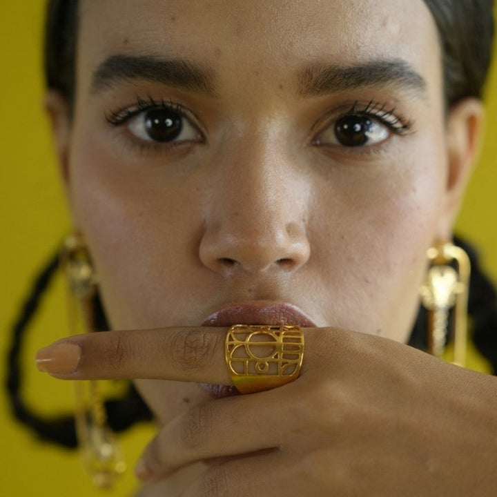 Close-up of woman with dark hair wearing gold geometric statement ring and matching earrings against yellow background