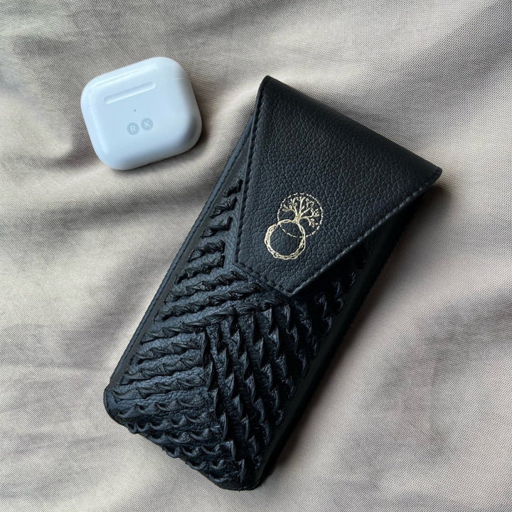 Black upcycled leather iPhone sling with woven design and gold tree logo, next to white wireless earbuds case