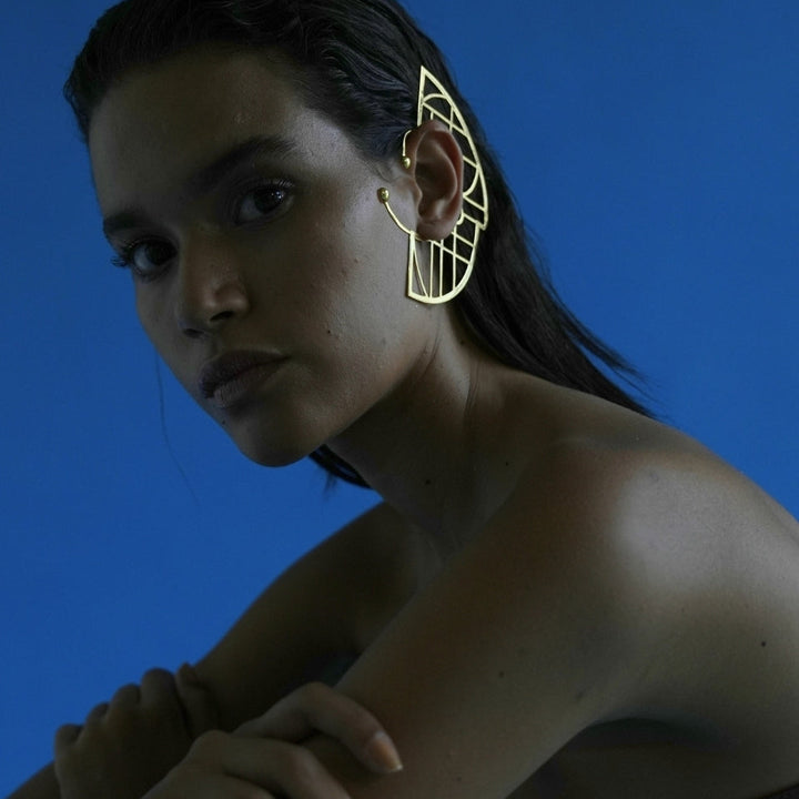 Model wearing an adjustable gold metal ear cuff with geometric design against a blue background