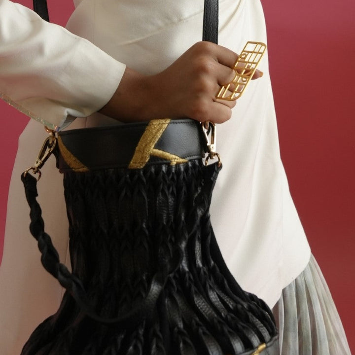 Hand holding gold geometric metal ring with black leather and woven handbag against maroon background