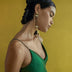 Woman with braided hair wearing a green satin dress and oversized geometric gold earrings against a mustard yellow background