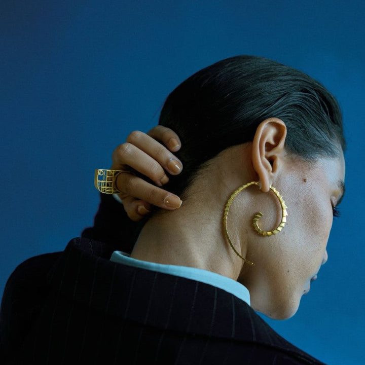 Side profile of woman with slicked black hair wearing large gold spiral earrings and a gold ring against blue background