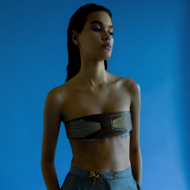 Model wearing a timeless tan leather upcycled waistbelt with golden embroidery and high-waisted blue pants against a blue background