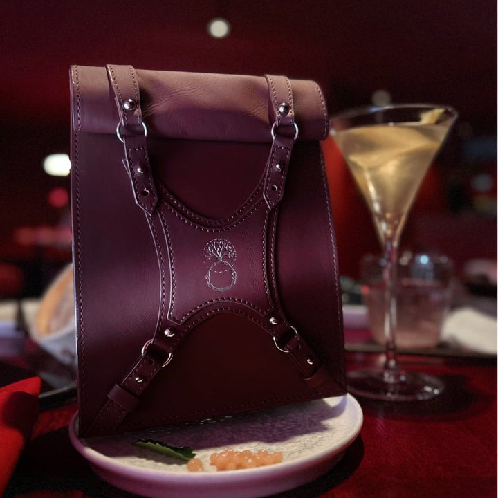 Dark red leather handbag with buckles on a plate beside a martini glass on red table