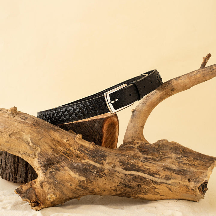 Black woven leather belt with silver buckle displayed on rustic wooden logs and beige sand