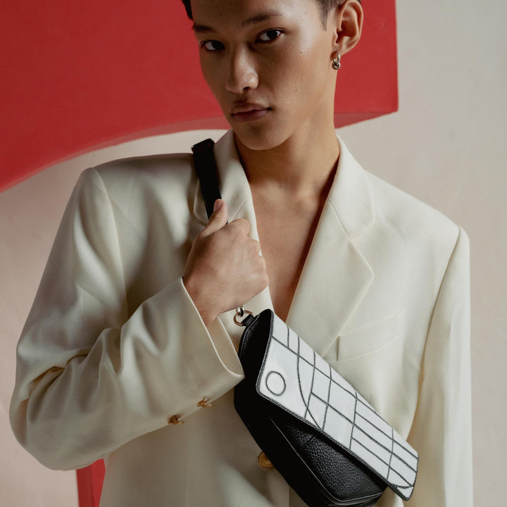 Stylish young man wearing cream blazer and holding black and white patterned crossbody bag against red and beige backdrop