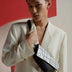 Stylish young man wearing cream blazer and holding black and white patterned crossbody bag against red and beige backdrop