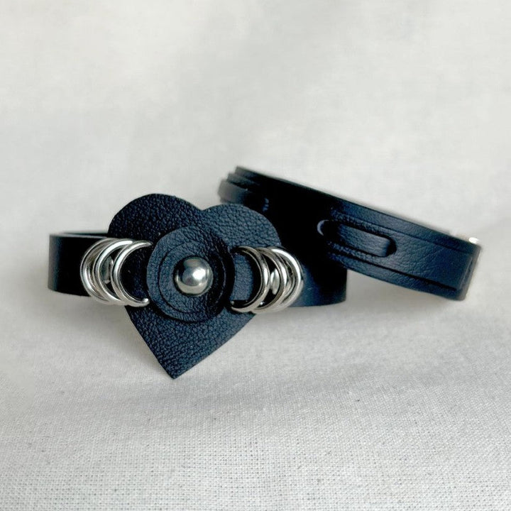 Two black leather cuff bracelets, one with heart-shaped centerpiece and silver rings, on light fabric