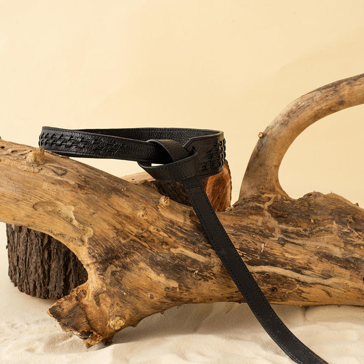 Black leather belt with woven details draped over natural driftwood on sand against beige background