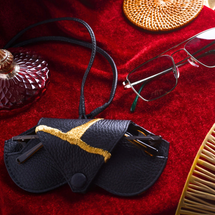Black leather eyeglass case with gold detail, eyeglasses, rattan coaster, and vintage perfume bottle on red velvet