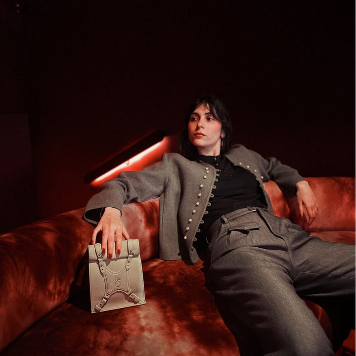 Woman in gray jacket and pants lounging on red velvet sofa holding beige handbag in dimly lit room