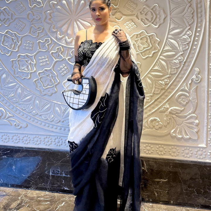 Woman wearing black and white saree with floral embroidery, holding crescent-shaped handbag, standing by a decorative wall