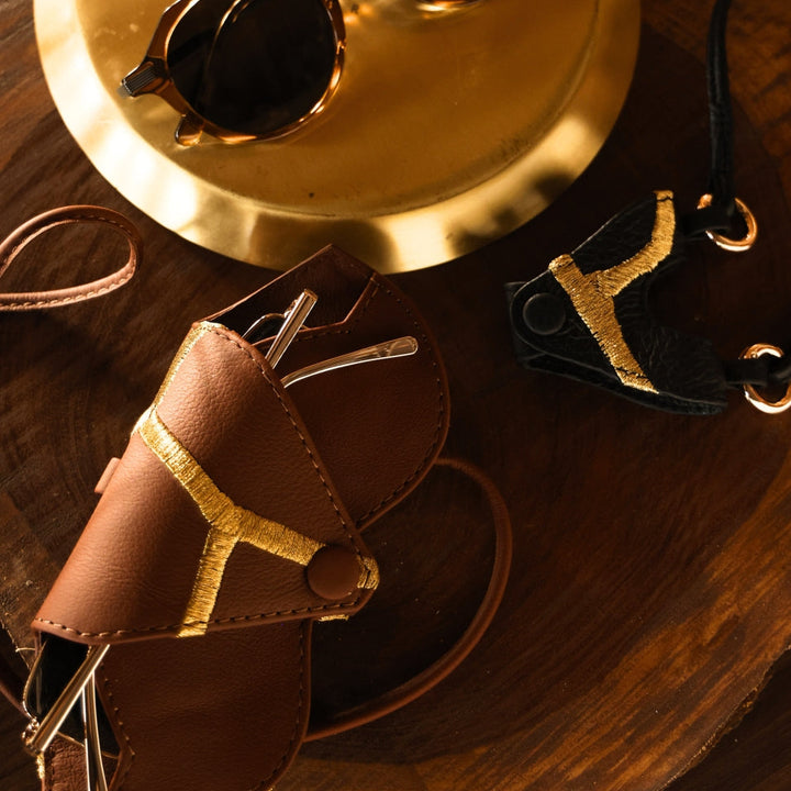 Brown and black leather eyeglass cases with gold embroidery, sunglasses on gold plate, wooden surface