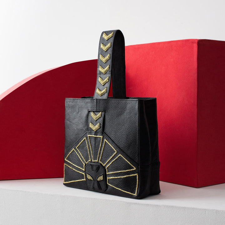Black leather tote bag embossed with gold geometric patterns and arrows handle, against red and white background