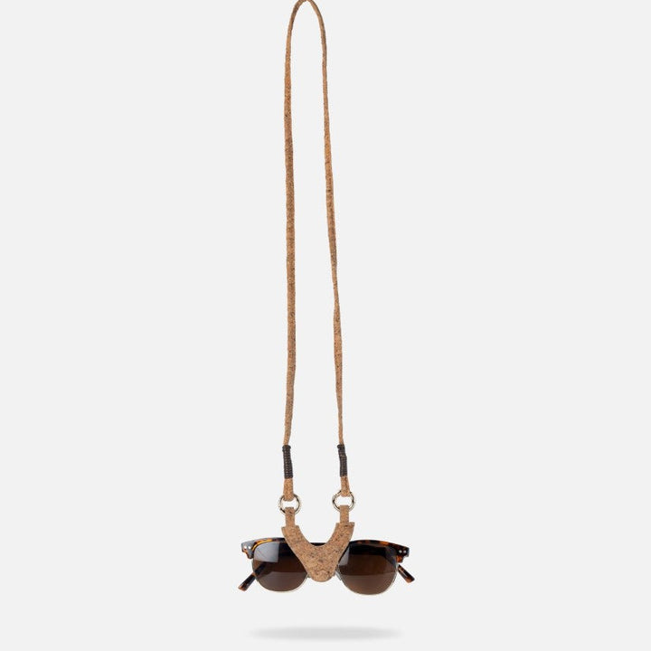Sunglasses with dark lenses held by a handcrafted cork and leather strap hanging against a white background