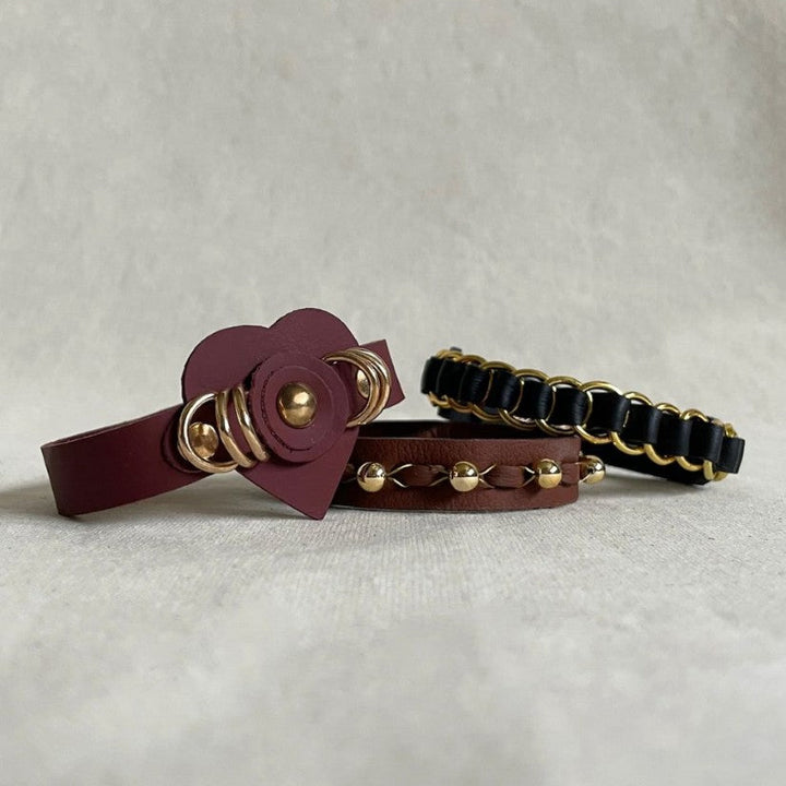 Three handcrafted leather bracelets in burgundy and black with gold rings and studs on a neutral surface