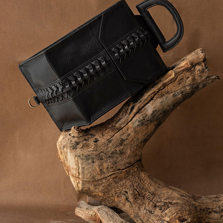 Black leather handbag with woven detail displayed on rustic wooden branch and beige fabric