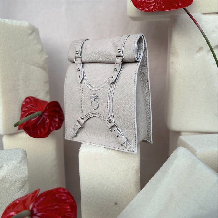Beige leather handbag with buckle straps, displayed among cream foam blocks and red anthurium flowers