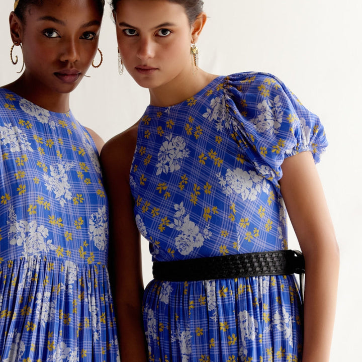 Two women wearing blue floral plaid dresses with puffed sleeves and black belt, posing against white background