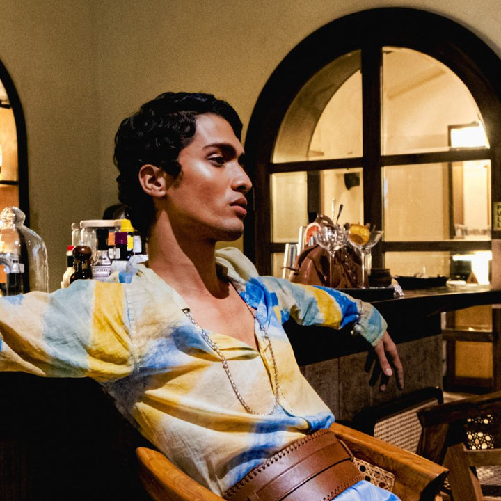 Man wearing a colorful tie-dye shirt and a wide tan leather waist belt, seated in a wooden chair indoors
