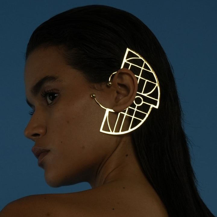 Profile of woman with sleek dark hair wearing large gold geometric metal ear cuff against blue background