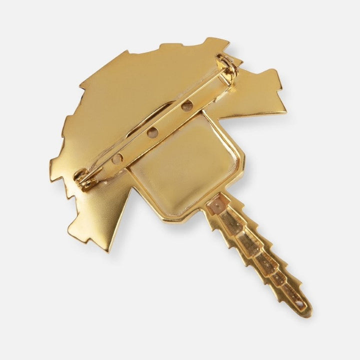 Back view of a gold metal brooch shaped like a key with a pin fastening on white background