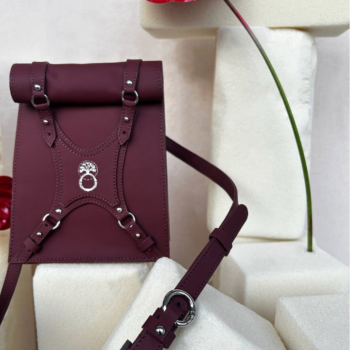 Maroon leather backpack with silver hardware and a rolled top, displayed on white foam blocks with a red anthurium flower