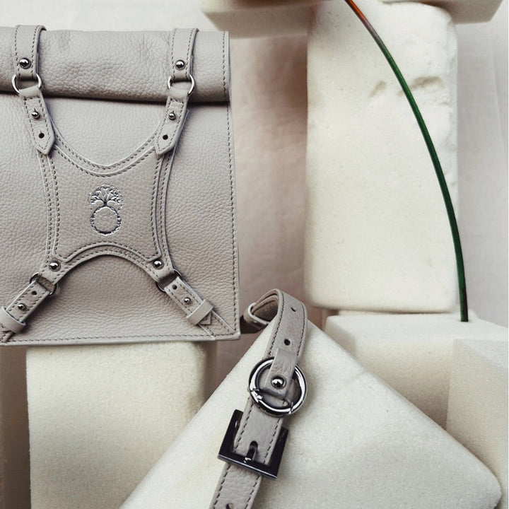 Minimalist beige leather handbag with silver buckle and embossed logo, displayed on white foam blocks with red flower