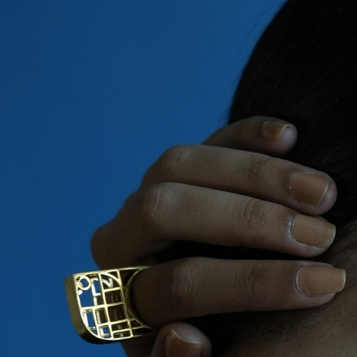 Hand wearing a large gold geometric metal ring against a solid blue background