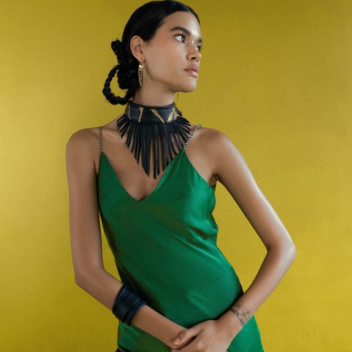 Woman in green dress wearing black fringe choker, gold earrings, and black bracelets against yellow background