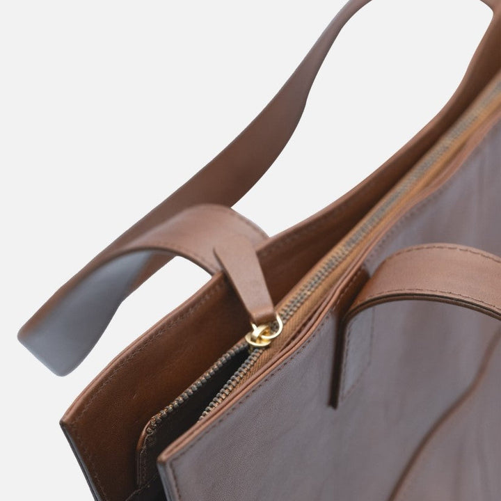 Close-up of brown leather tote bag with gold zipper and sturdy handles on white background