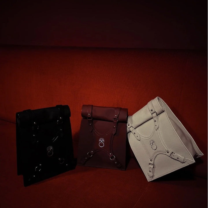 Three leather roll-top backpacks in black, brown, and white resting on a red fabric sofa