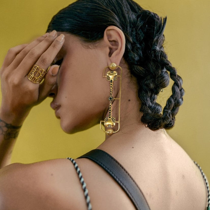 Close-up of a woman with braided hair wearing a large, intricate gold earring and a gold ring against a yellow background