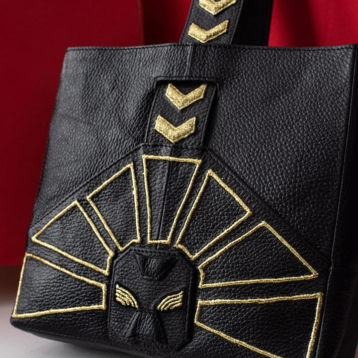 Black leather tote bag with gold geometric embroidery and bold strap design against red background