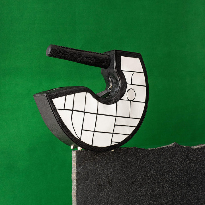 Black and white crescent-shaped handbag with geometric pattern on gray stone against green background