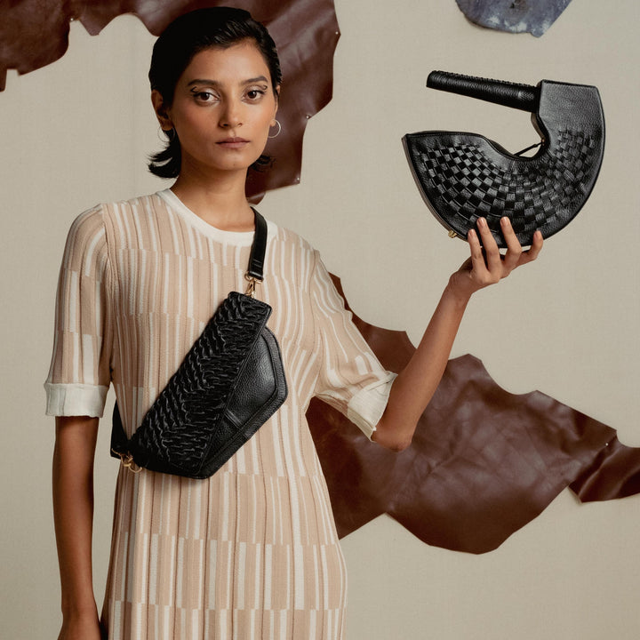 Woman in beige striped dress holding and wearing black woven leather bags against neutral backdrop with leather pieces