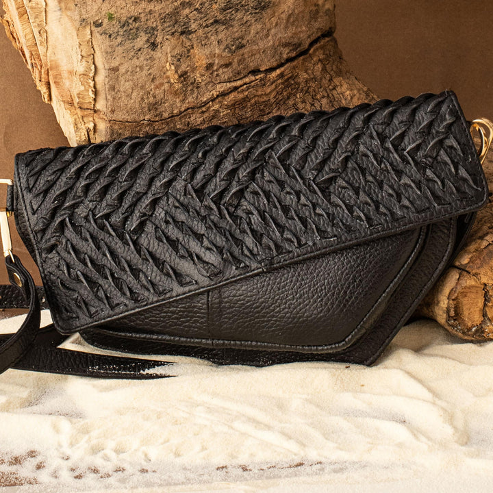 Black woven leather crossbody bag with detachable belt, displayed on white sand and rustic wood