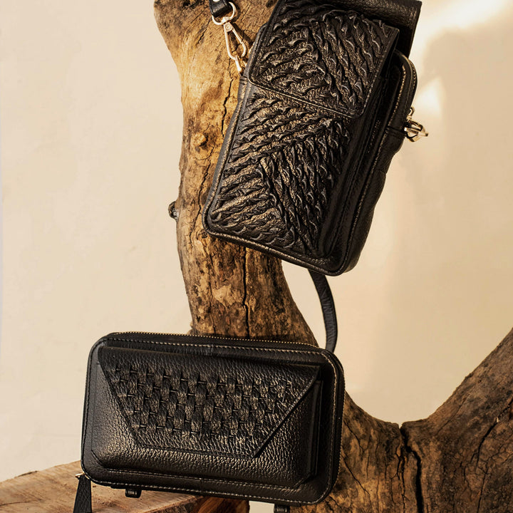 Two black textured leather crossbody bags displayed on natural wooden branches